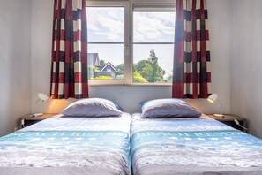 Ein Doppelbett mit einem Bettbezug im Strandstil, zwei Nachttischlampen und ein Fenster mit Vorhängen.