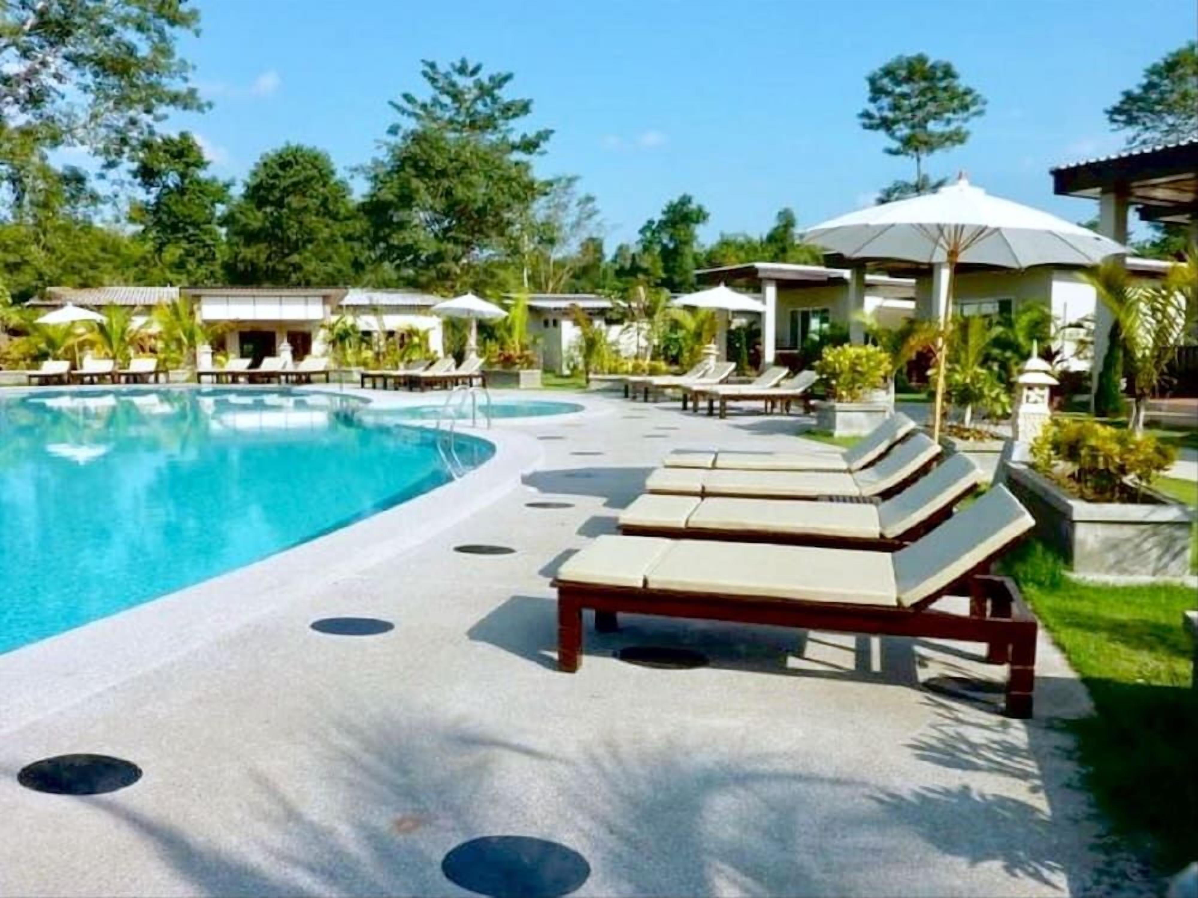 A pool area with lounge chairs and umbrellas, surrounded by greenery and buildings.