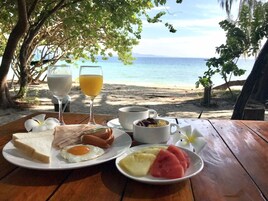 A breakfast table with a plate of toast, egg, and fruit, a glass of orange juice, and a cup of coffee, set on a wooden table with a view of the beach and ocean.