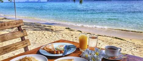 A wooden table with breakfast items including toast, fruit, and a beverage, set against a beach backdrop with clear skies.
