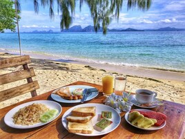 A wooden table with breakfast items including toast, fruit, and a beverage, set against a beach backdrop with clear skies.