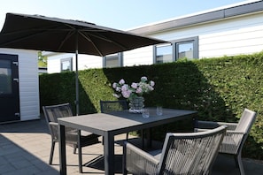 En udendørs terrasse med et bord, stole og en stor parasol, med en hæk i baggrunden.