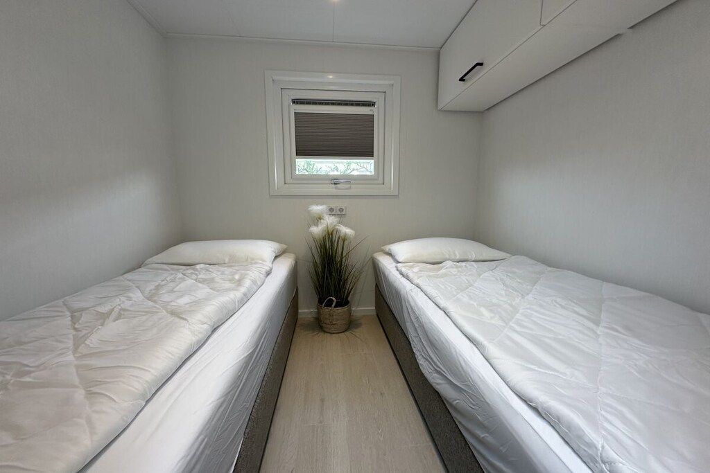 Une petite chambre propre, avec deux lits simples, une fenêtre avec des stores, une plante en pot et une bouche d’aération au plafond.