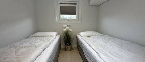 A small, clean room with two single beds, a window with blinds, a potted plant, and a ceiling vent.