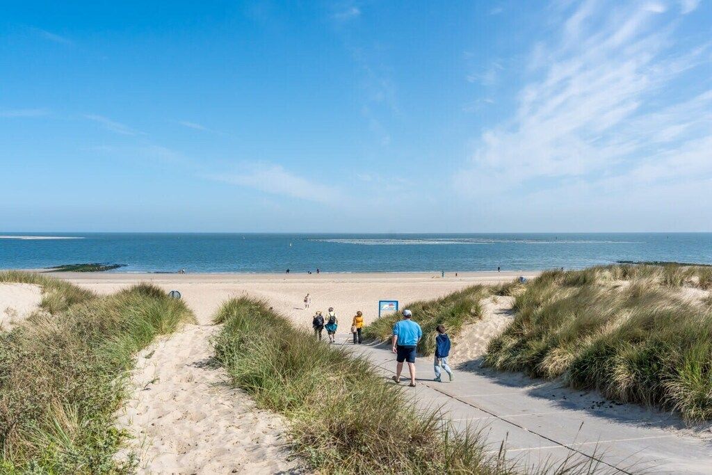 Une plage avec un ciel dégagé, un sentier sablonneux menant à l’eau, et des gens qui se promènent.