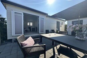 Ein Terrassenbereich im Freien mit Tisch und Stühlen, eine Glastür zum Innenbereich und ein überdachter Bereich ebenfalls mit Tisch und Stühlen.