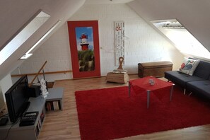 Ein Loft-Zimmer mit rotem Teppich, einem Fernseher, einem Bücherregal, einem roten Tisch, einer Couch und einem Gemälde eines Leuchtturms.