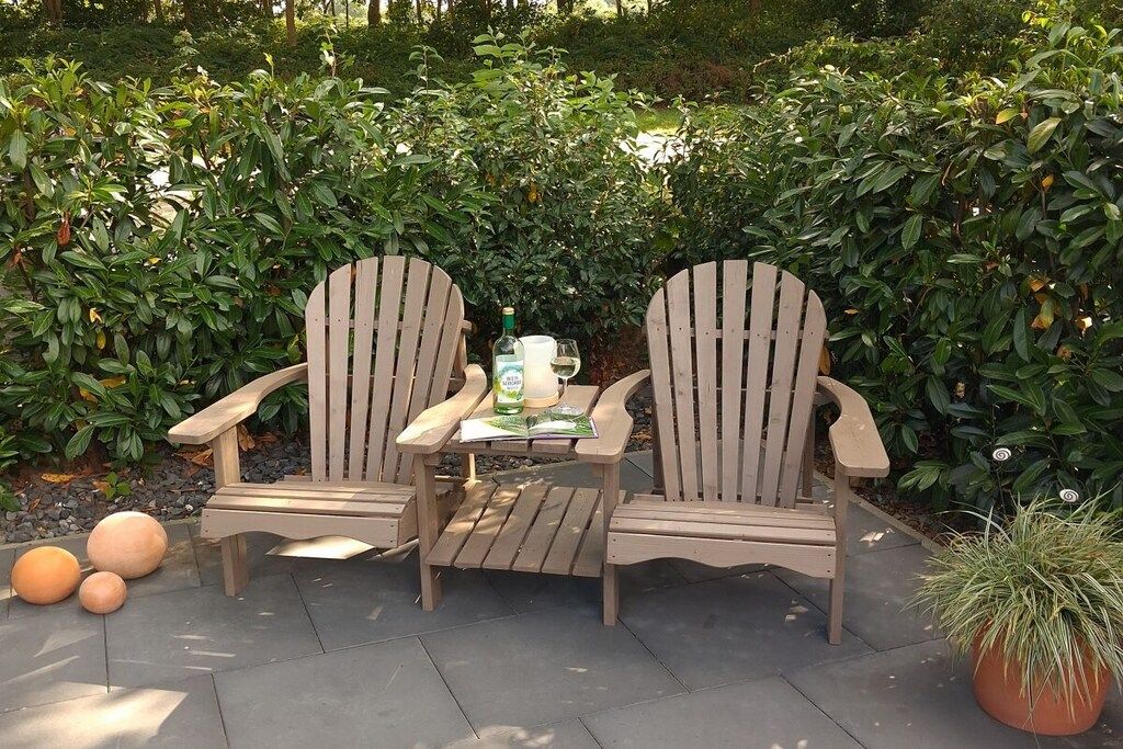 Two wooden Adirondack chairs with a table between them, placed on a paved patio surrounded by greenery.