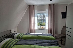 Ein Schlafzimmer mit zwei Betten, einem Fenster mit Vorhängen, einem Sessel und einem an der Wand montierten Fernseher.