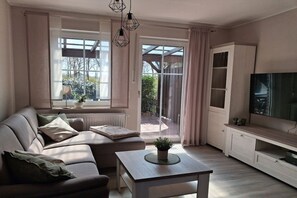 Sala de estar com sofá, mesa de centro, televisão e porta de vidro deslizante que dá para área externa.