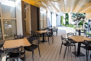 Uma área de café ao ar livre com mesas de madeira e cadeiras pretas, grandes janelas e um espaço coberto para sentar.