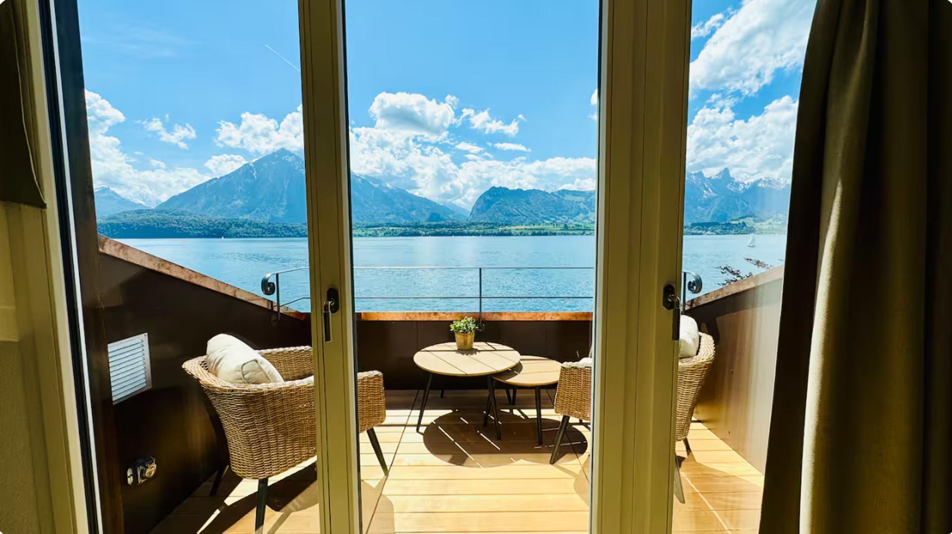 A balcony with wicker furniture, a small table, and a view of a lake and mountains.