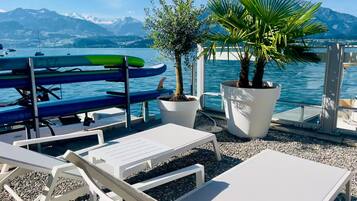 Una terraza con sillones blancos, una palmera pequeña y una fila de barcos.
