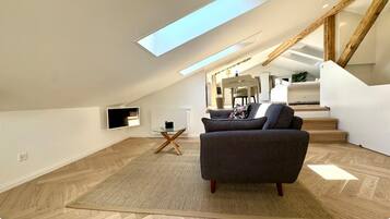 Un loft moderno con techo alto, tragaluz, sofá de color gris oscuro, una pequeña mesa de vidrio y una escalera que sube a un nivel superior.