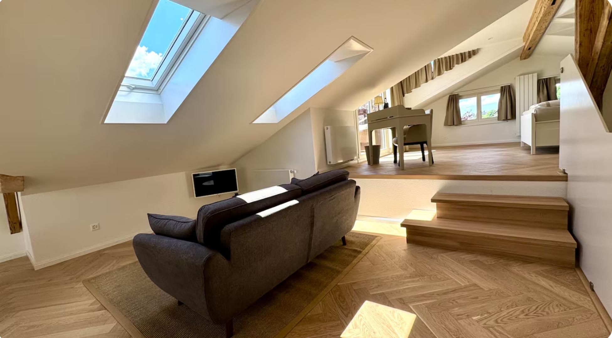 Un loft espacioso con techo inclinado, sofá integrado, zona de comedor y escalera que lleva a un nivel superior.