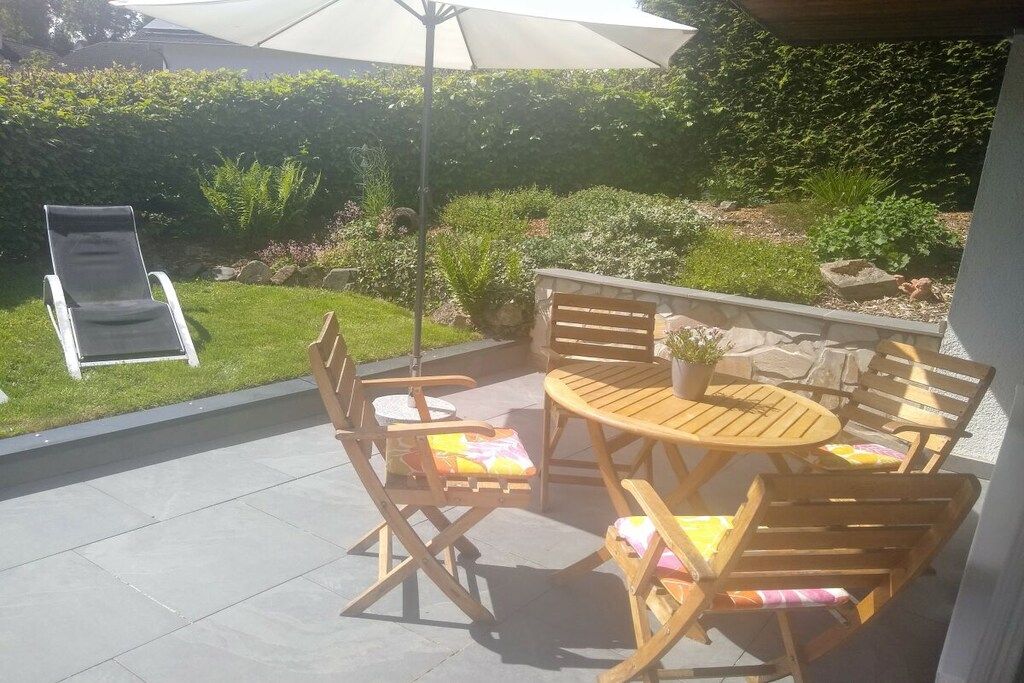 Een terras buiten met een ronde houten tafel en stoelen, een opklapbare stoel en een bloempot.