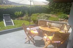 Een terras buiten met een ronde houten tafel en stoelen, een opklapbare stoel en een bloempot.
