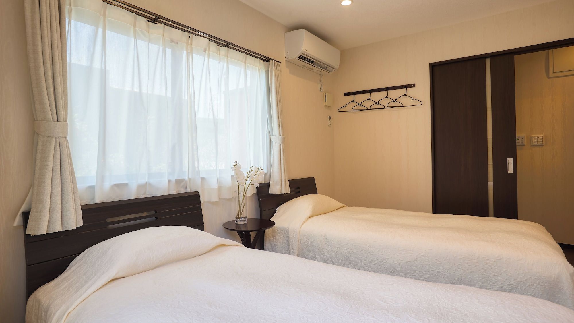 A hotel room with two beds, a window with sheer curtains, an air conditioning unit, a bedside table with a vase of flowers, and a coat rack.