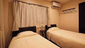 A room with two beds, a small table, a hanging clothes rack, and an air conditioning unit.
