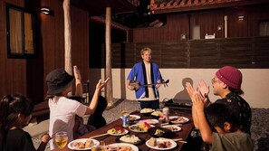 Un grupo de personas sentadas en una mesa al aire libre con comida y bebidas, una persona tocando la guitarra, y una pared de madera con un cuadro enmarcado.