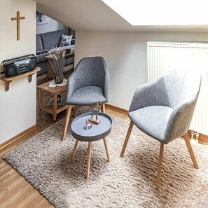 Un salón acogedor con dos sillones grises, una mesita redonda, una mesita auxiliar de madera, una radio y una cruz en la pared.