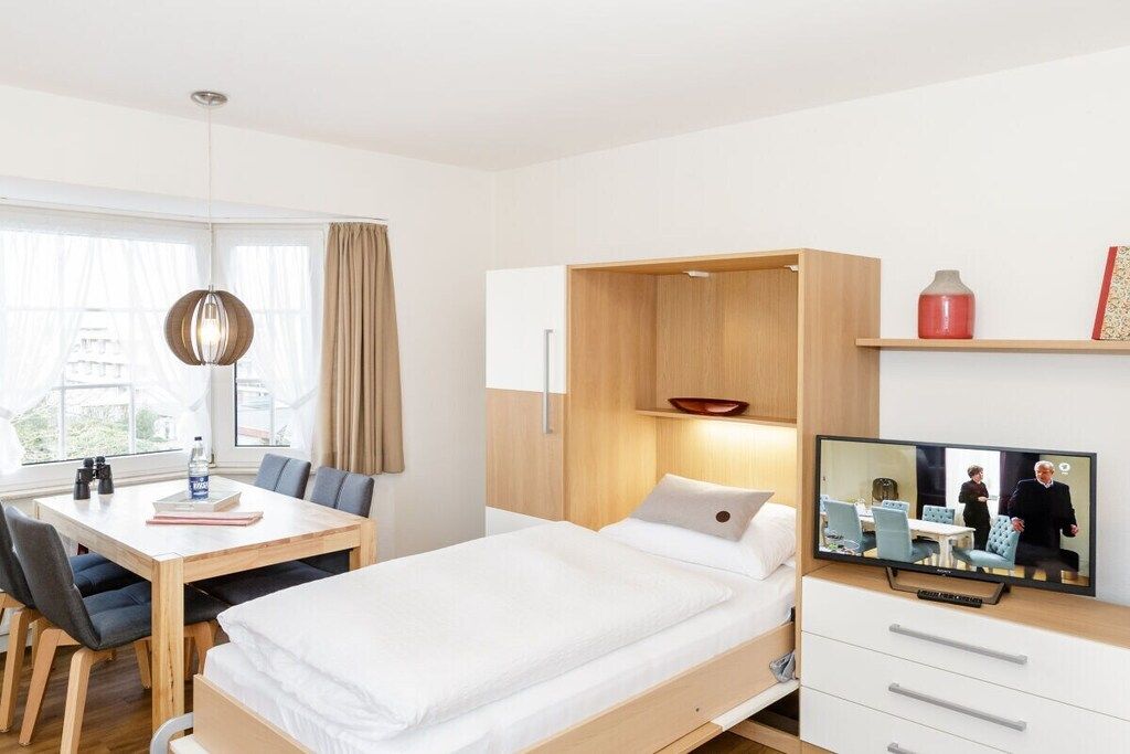 A modern hotel room with a bed, a desk with chairs, a television, and a window with curtains.