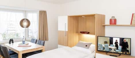 A modern hotel room with a bed, a desk with chairs, a television, and a window with curtains.