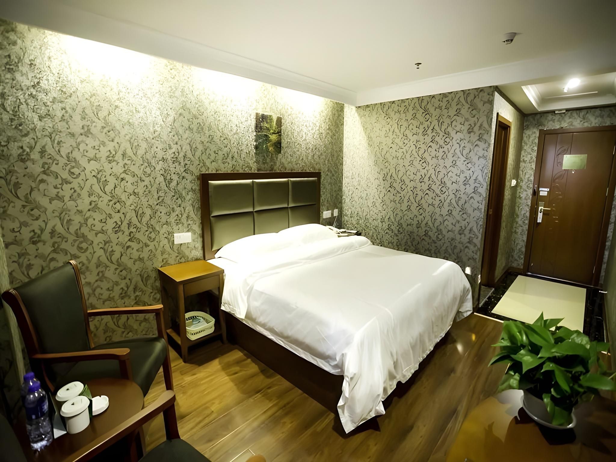 A hotel room with a bed, a chair, a nightstand, a potted plant, and a wooden floor.
