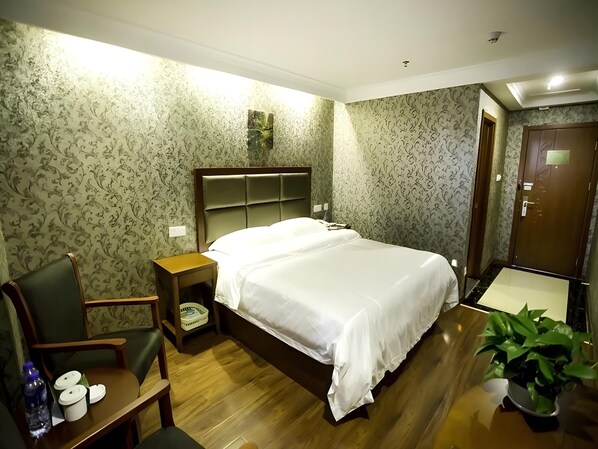 A hotel room with a bed, a chair, a nightstand, a potted plant, and a wooden floor.