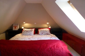 Een slaapkamer met schuine plafonds, een bed met rode beddengoed, twee bedlampjes en een klein schilderij aan de muur.