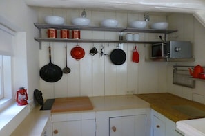 Een keuken met witte kastjes, een houten aanrecht, hangende pannen, een magnetron en planken met kommen en potten.