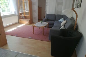 Ein Wohnzimmer mit einer Couch, einem kleinen Tisch mit Pflanze, einem schwarzen Sessel, einem Holzschrank und einem Fenster.