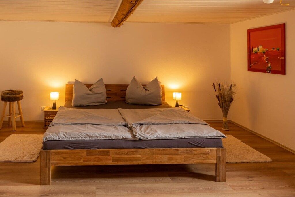 Un dormitorio con una cama de madera, dos lámparas de noche, una mesita de noche, un cuadro en la pared y un florero con plantas secas.