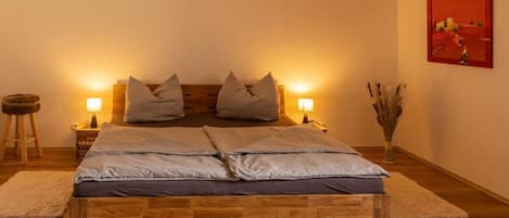 Un dormitorio con una cama de madera, dos lámparas de noche, una mesita de noche, un cuadro en la pared y un florero con plantas secas.