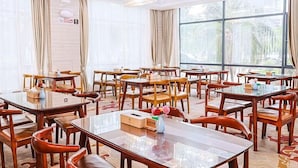 Un comedor de hotel con varias mesas y sillas, ventanas amplias con cortinas y un candelabro.