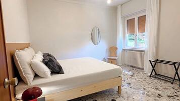 Una camera da letto con un letto, un comodino, uno specchio, un termosifone, una finestra con tende e una lampada a sospensione.