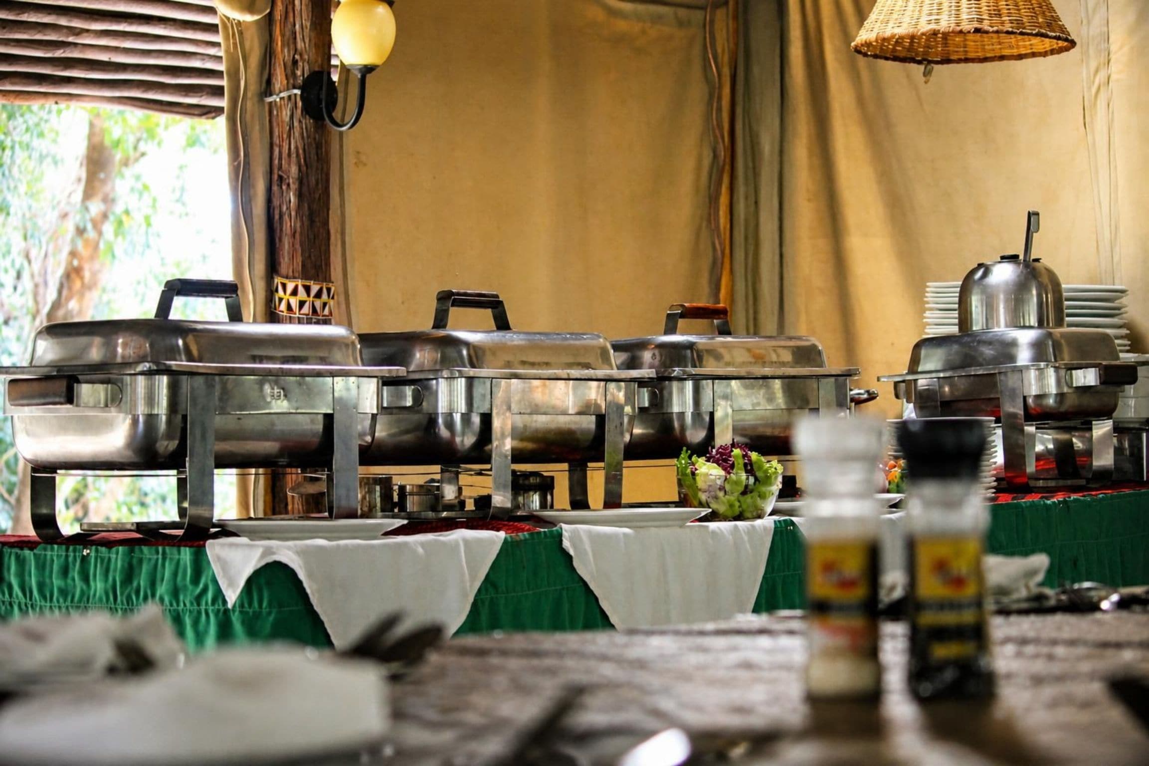 A buffet setup with multiple stainless steel chafing dishes, a table with a green tablecloth, and a wicker hanging light fixture.