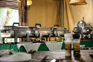 A buffet setup with multiple stainless steel chafing dishes, a table with a green tablecloth, and a wicker hanging light fixture.