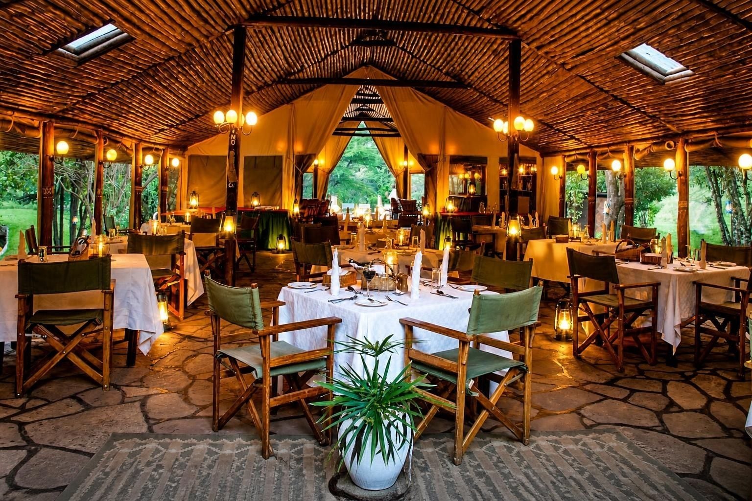 Un espacio de comedor al aire libre con mesas dispuestas para dos, rodeado de sillas de madera y una estructura de techo de paja.