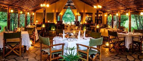 Un espacio de comedor al aire libre con mesas dispuestas para dos, rodeado de sillas de madera y una estructura de techo de paja.