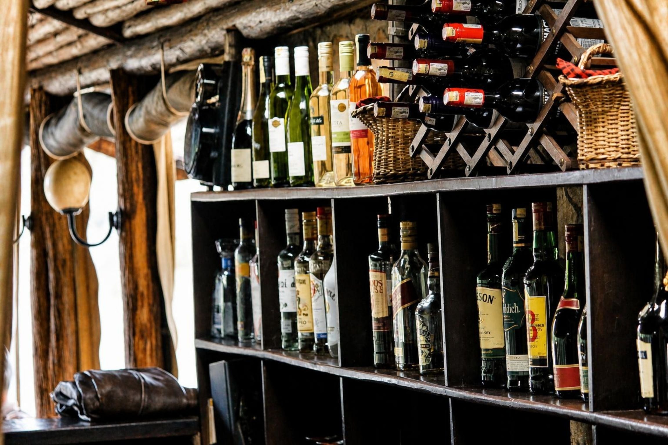 A bar area with shelves stocked with various bottles of alcohol, a leather chair, and a rustic wooden ceiling.