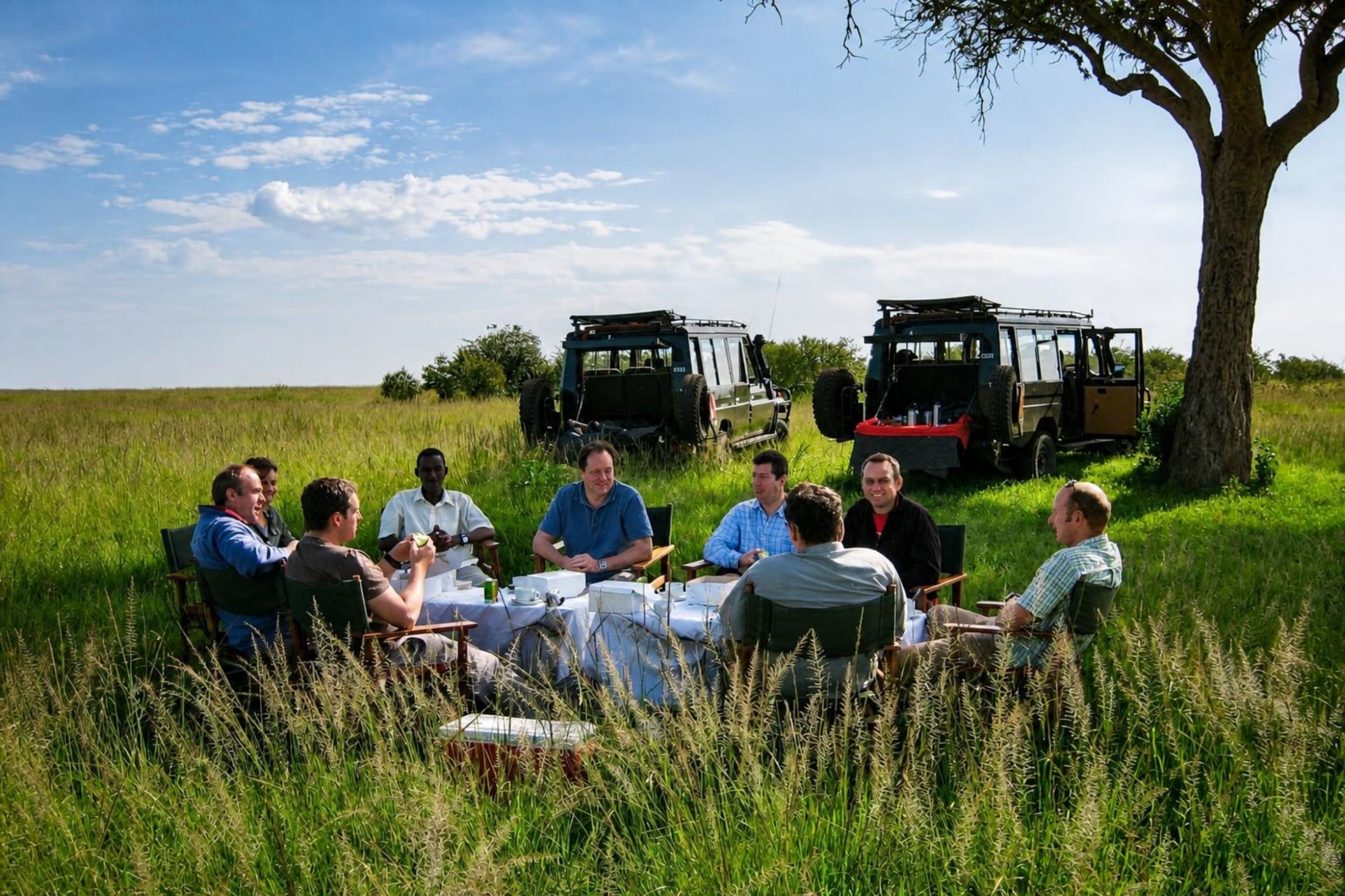 Un grupo de personas sentadas alrededor de una mesa al aire libre, con vehículos al fondo.