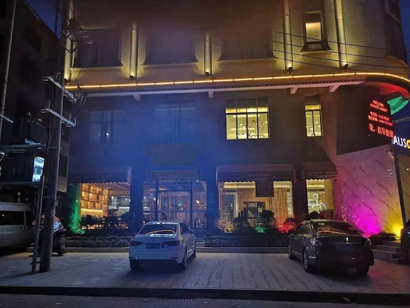 A hotel entrance with a car parked in front, a signboard with text, and a building facade with windows.