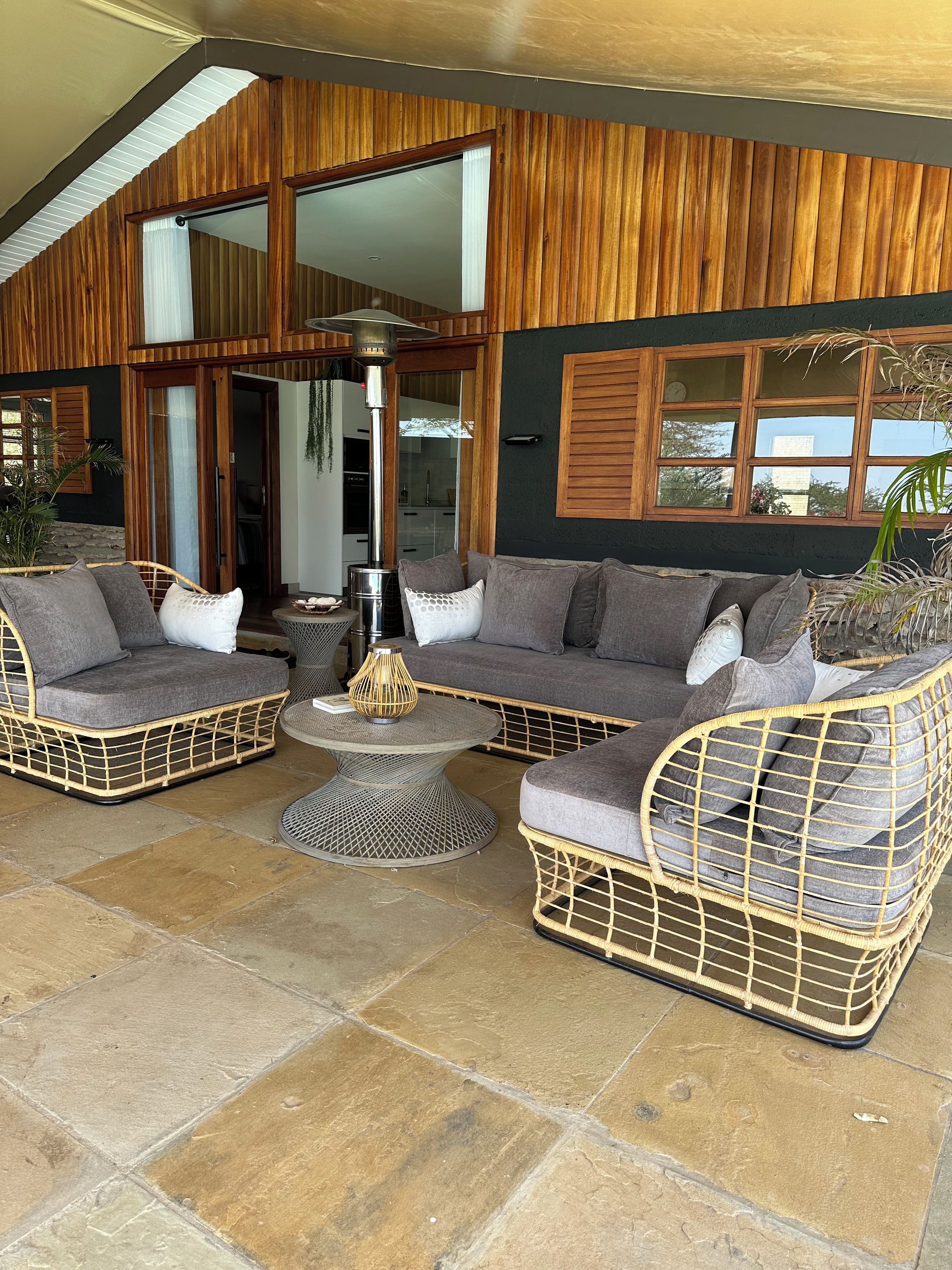 A cozy outdoor seating area with wicker furniture and a central table, set against a backdrop of wooden paneling and large windows.
