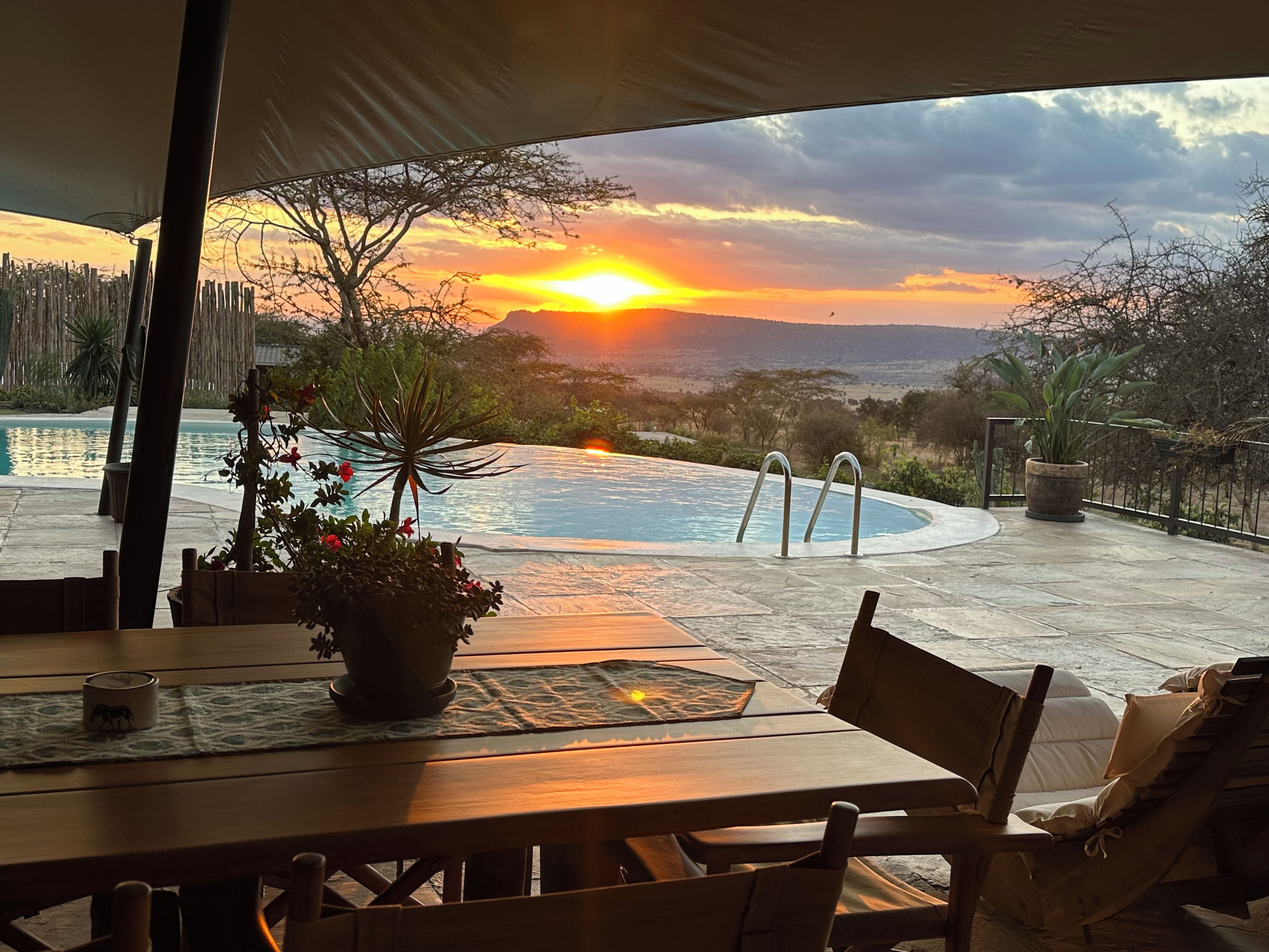 A poolside area with a view of the sunset, a wooden table, and lounge chairs.