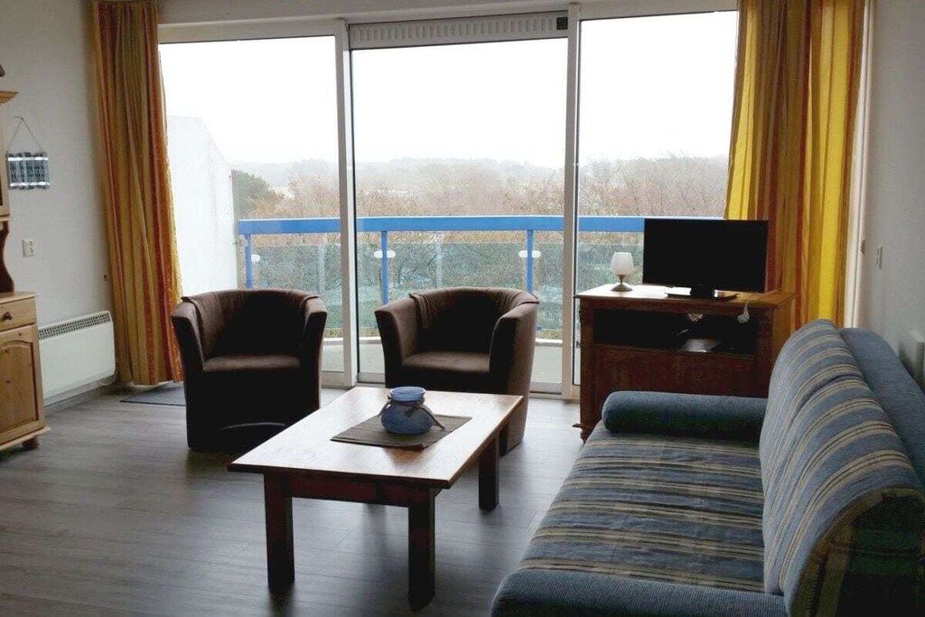 A living room with a sofa, two armchairs, a coffee table, a TV on a stand, and a view through a large window.
