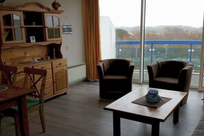 Ein Wohnzimmer mit Holzschrank, zwei Sesseln, einem Esstisch und Blick durch ein großes Fenster.