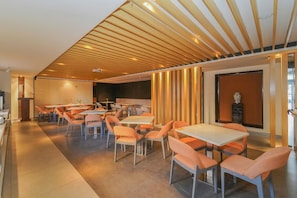 Uma sala de jantar moderna com várias mesas e cadeiras, tábuas de madeira no teto e uma tela grande na parede.