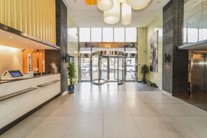 Área de recepção de hotel com um quiosque de check-in digital, uma porta giratória e um balcão de atendimento.
