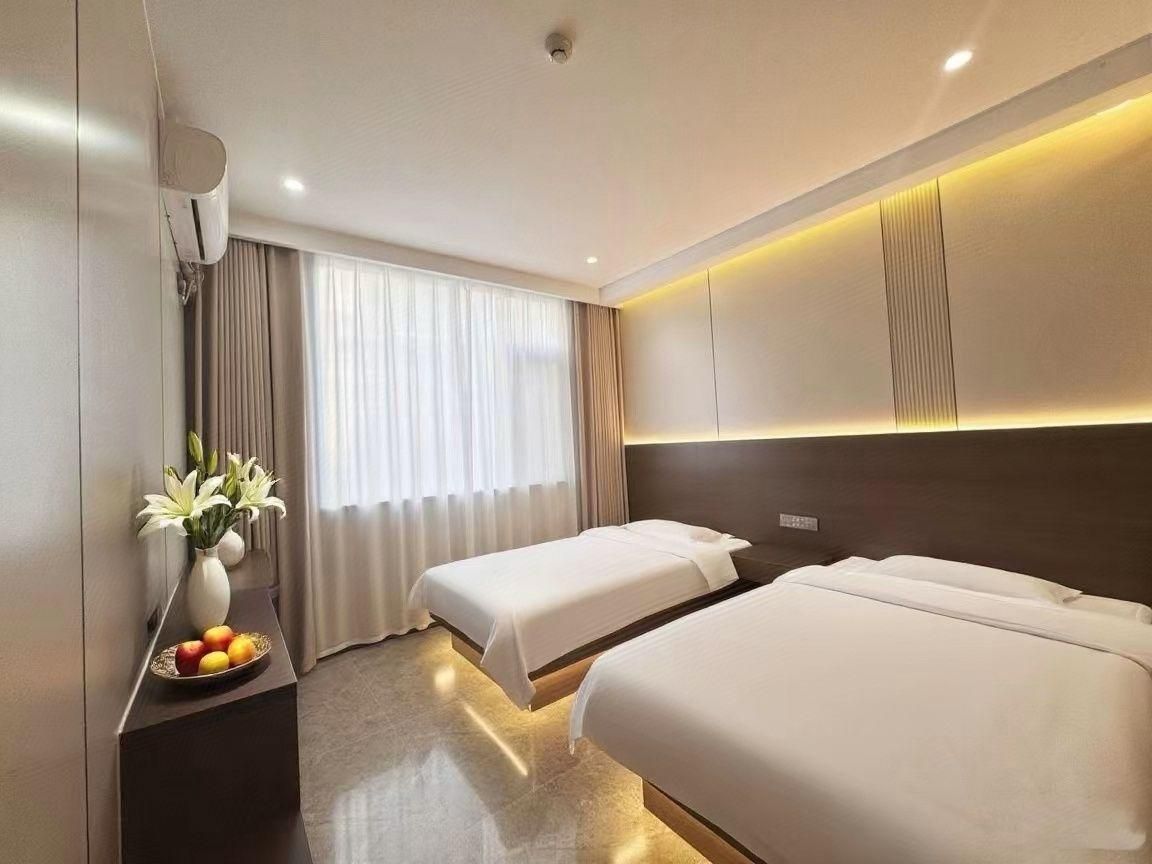 A hotel room with two beds, a bedside table with a vase of flowers, and a large window with curtains.